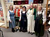 Fem kvinner i tradisjonelle drakter står foran hyller med tekstiler og varer.