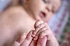 Nærbilde av babyhånd som holder en voksen finger, med et spedbarn i uskarp bakgrunn.