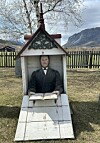 Lite kapellformet gravminne med skulptur og åpen bok på en gressplen foran fjell og gjerde.