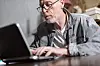 En eldre mann med briller sitter bøyd over en laptop på det som skal være et hjemmekontor.