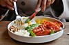 Person spiser med kniv og gaffel fra tallerken med røkt laks, egg, ost, nøtter, avokado og litt tomat.