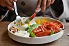 Person spiser med kniv og gaffel fra tallerken med røkt laks, egg, ost, nøtter, avokado og litt tomat.