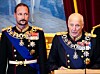Kronprins og konge på stortinget i dekorerte mørke paradeuniformer foran et blått bakteppe og røde gardiner.
