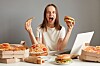 Smilende kvinne med pizzabit og burger ved et bord med pizza, burger og bærbar PC.