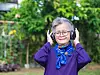 Eldre kvinne med briller, lilla stripet genser og blått skjerf bruker trådløse hodetelefoner i en grønn hage.