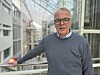 Bespectacled man in a grey jumper standing beside a railing in a glass atrium.