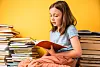 Skolejente leser en rød bok omgitt av stabler med bøker.