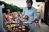 Far og sønn griller sammen på en terrasse, med grillmat i forgrunnen og familie i bakgrunnen.