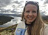 Erin Lindsay smiler foran en innsjø med skogkledde fjell og skyer.