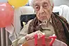 En eldre kvinne blåser ut lysene på en kake der tallene danner tallet 117, for å feire bursdagen sin. I bakgrunnen ses røde og gule ballonger.