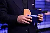 Programleder i NRK ikledd mørk dress holder et oransje kort med teksten «Debatten» i et studio-lignende miljø.